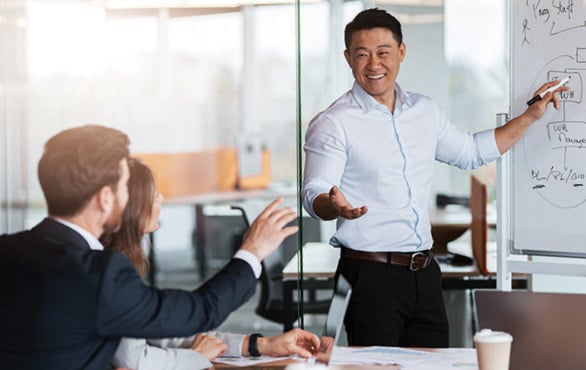 Professional presenting strategy to colleagues in a conference room while standing beside a whiteboard during a leadership meeting.