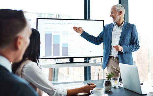 Senior executive delivering a presentation to a team using a large screen displaying business performance data.