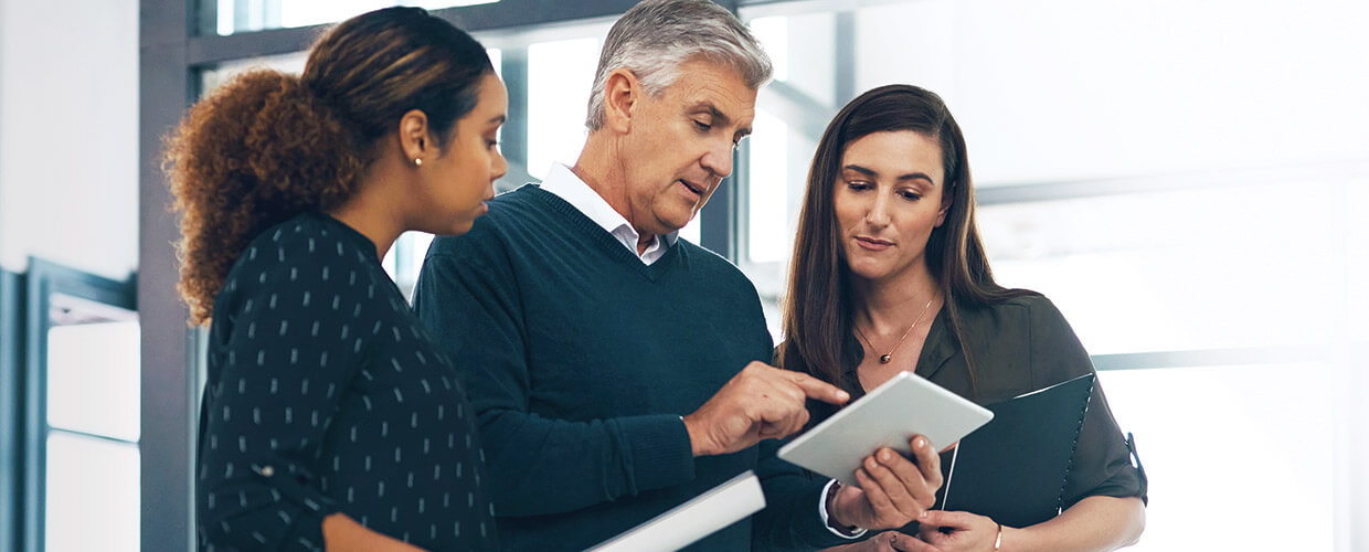 Three professionals collaborating around a tablet in a modern office, demonstrating workplace learning, coaching and knowledge sharing.