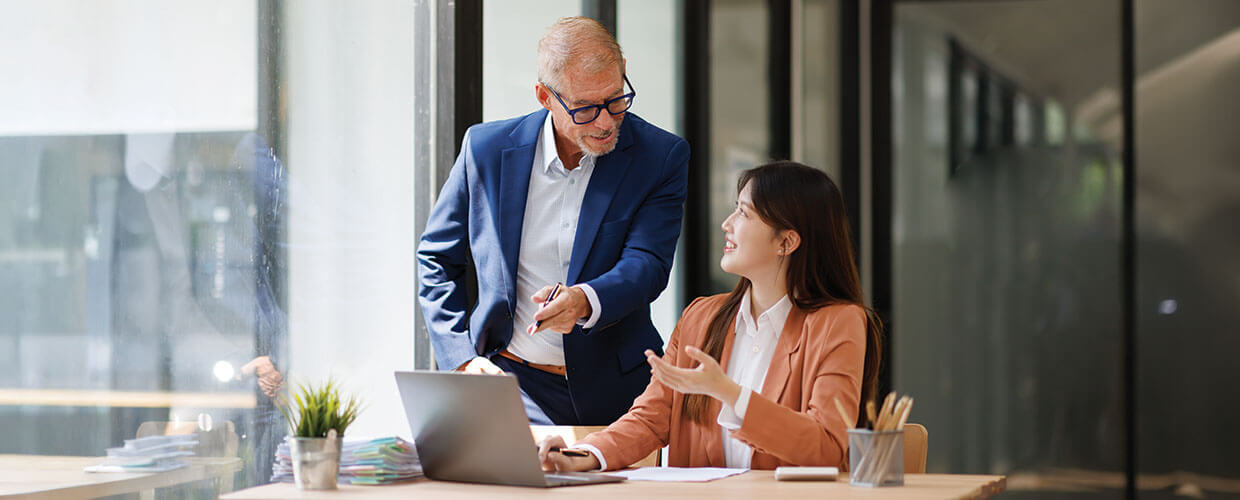 Senior business professional mentoring a younger colleague at a laptop in a modern office, demonstrating employee development and on-the-job learning.