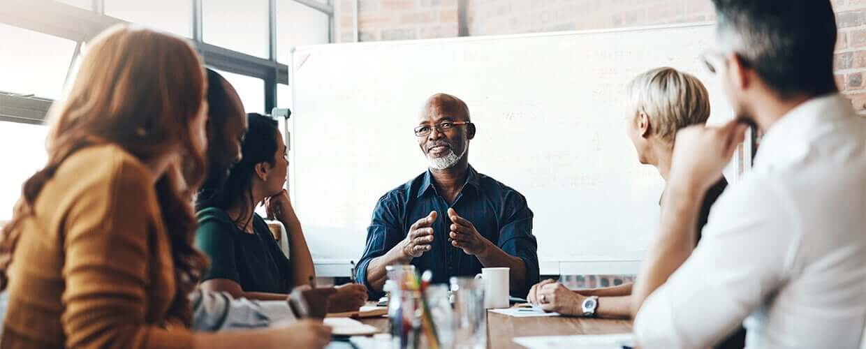 A business leader speaks to a diverse group of colleagues during a team meeting, explaining ideas in front of a whiteboard.