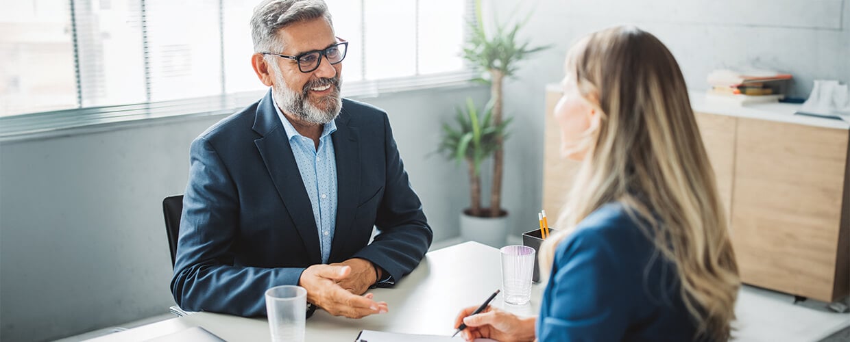 A business professional and a colleague sit across from each other in an office, engaged in a friendly discussion during a meeting or interview.