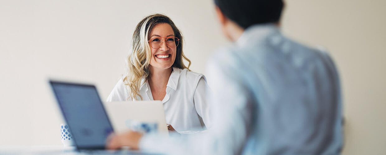 A smiling business professional meets with a colleague across a table, both using laptops during a discussion in a modern office setting.