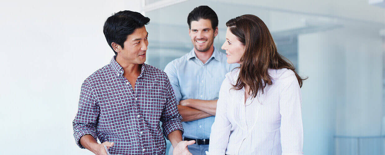 Professionals having a discussion in a modern office environment, with one man in a plaid shirt actively speaking, while a woman listens and a man stands in the background, smiling