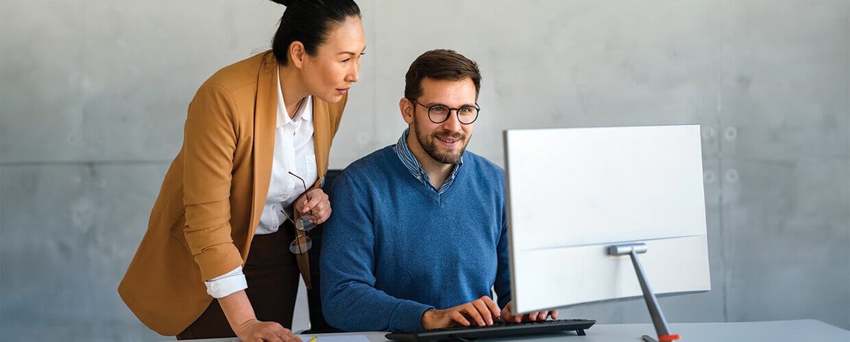 Manager coaching an employee at a computer while reviewing work and adapting to changing business priorities.